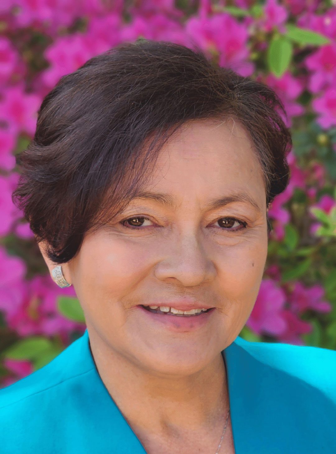 A woman with short dark hair and a turquoise jacket smiles in front of a backdrop of bright pink flowers.